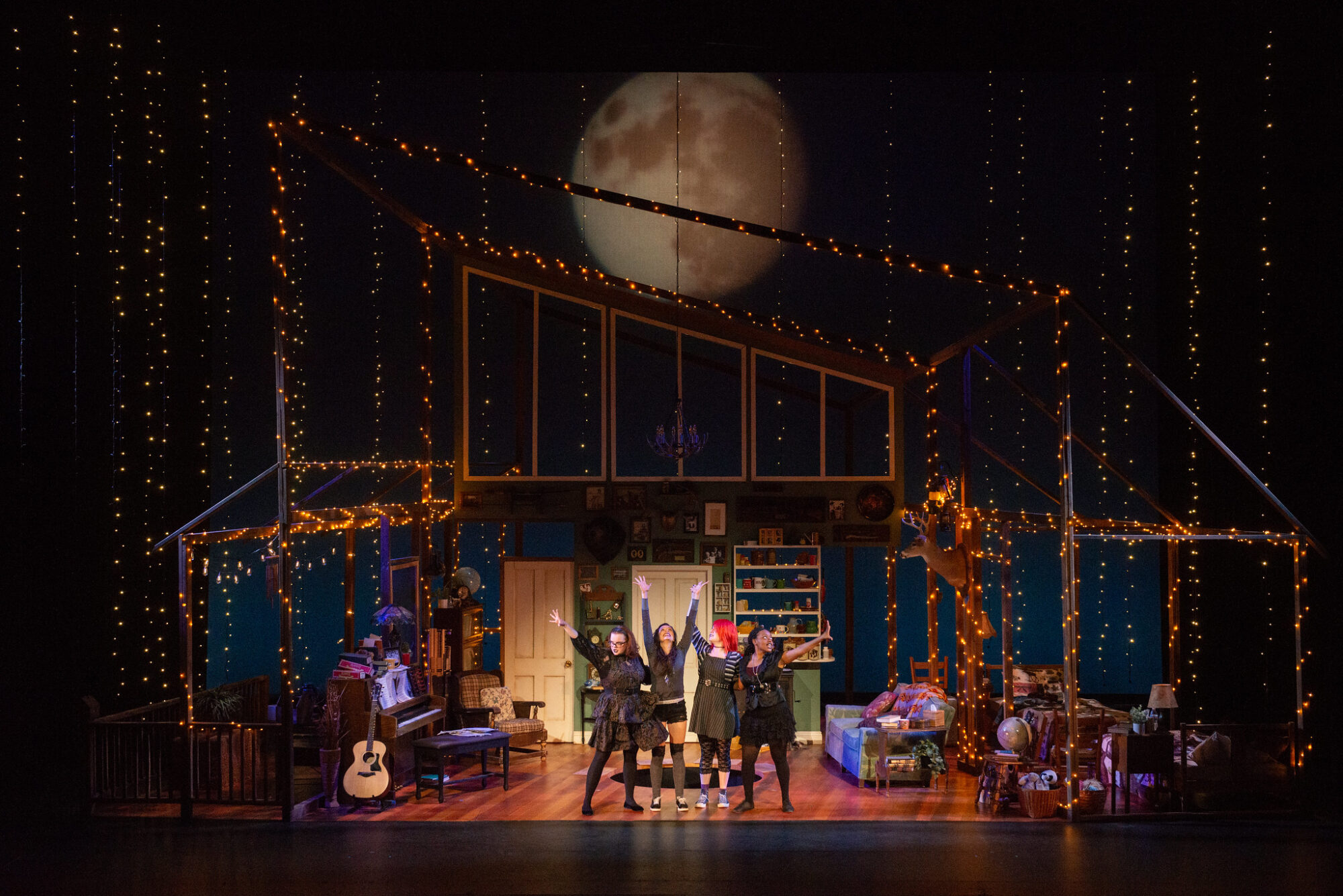 A large set that looks like a living room, covered in white twinkle lights, with a large projection of the moon in the background, four actors center stage with their arms raised in the air.